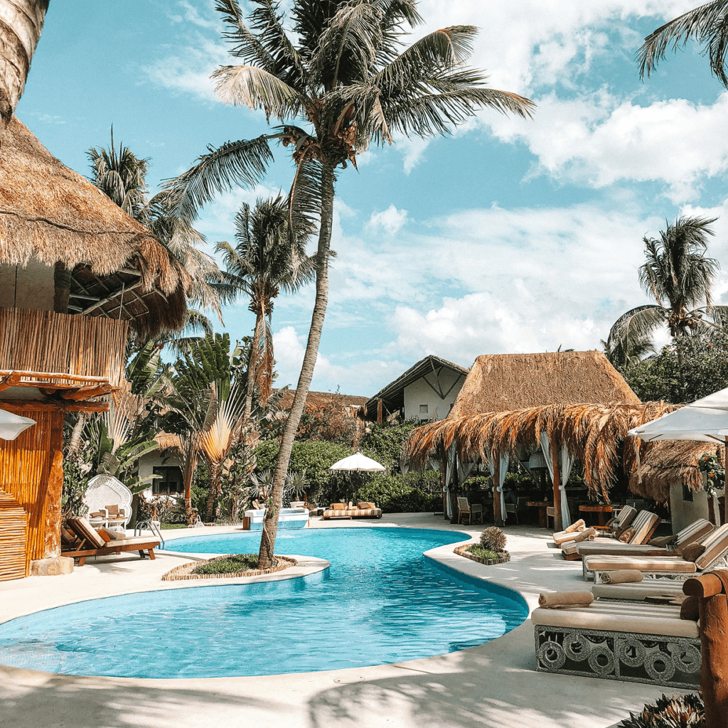 Tropical resort pool surrounded by palm trees and thatched huts under a bright sky.