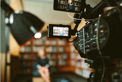 Video camera filming a person seated in a library setting.